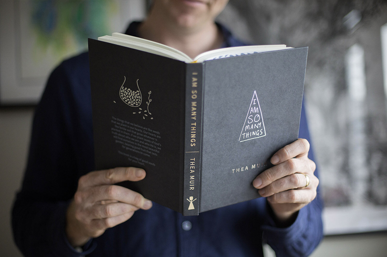 'I Am So Many Things' by Thea Muir, charcoal grey cloth edition, held open showing the hand drawn seed motif