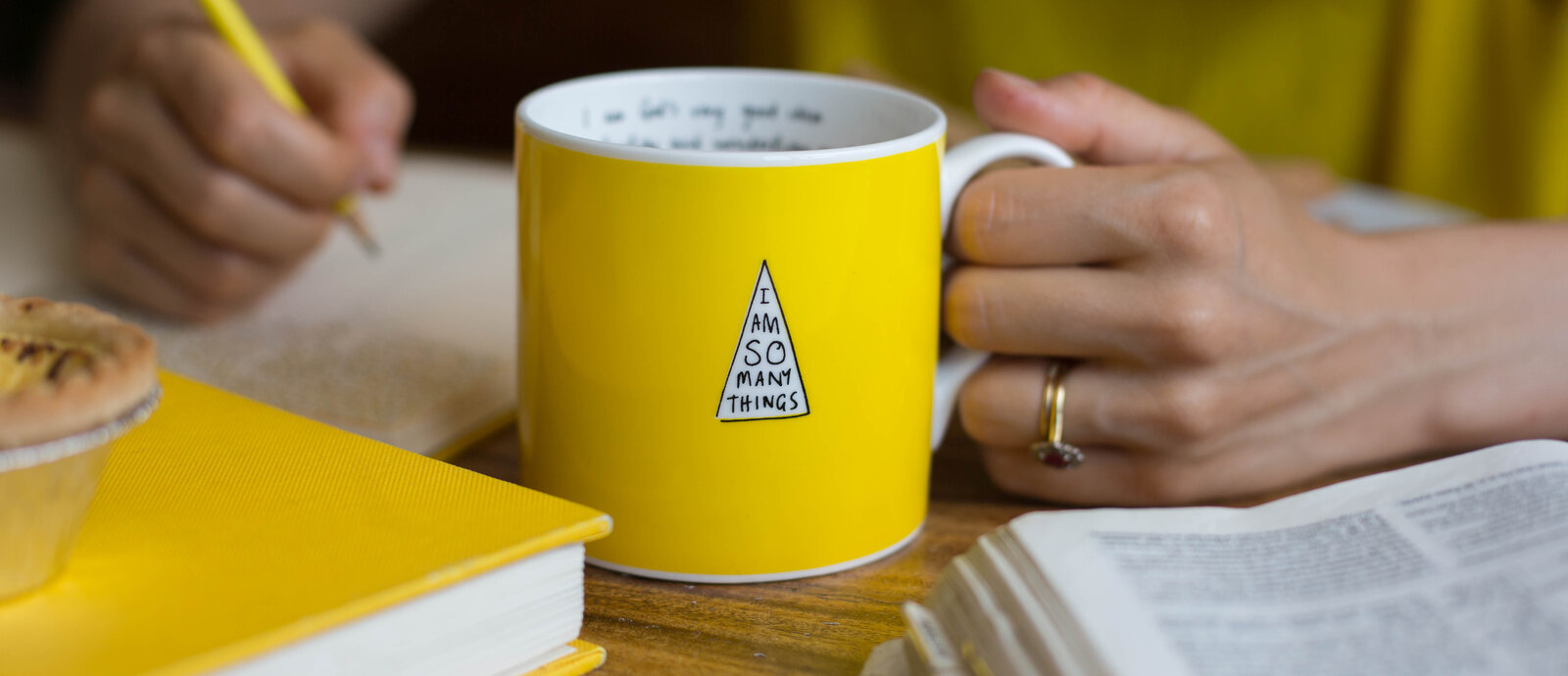 Yellow 'I Am So Many Things' mug held on a desk beside the book and a notebook
