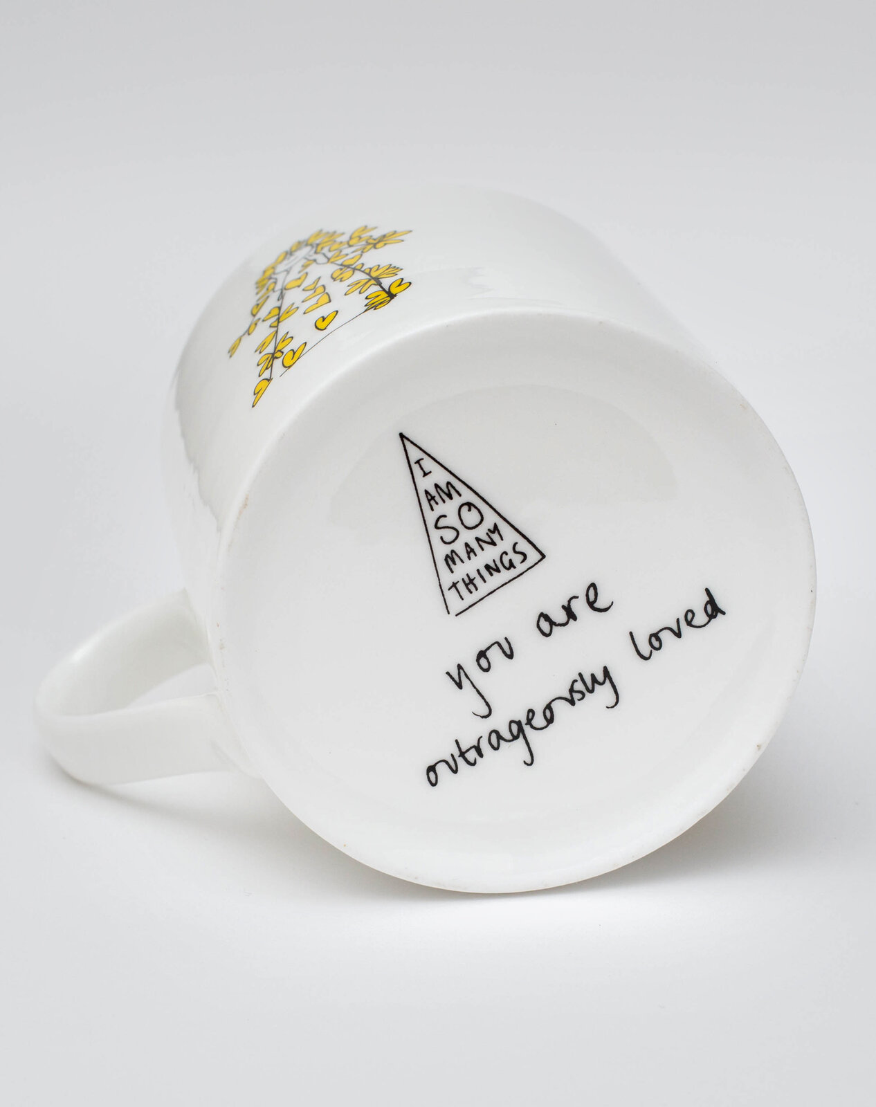 Base of an 'I Am So Many Things' mug, reading 'you are outrageously loved' beneath the book's triangle mark