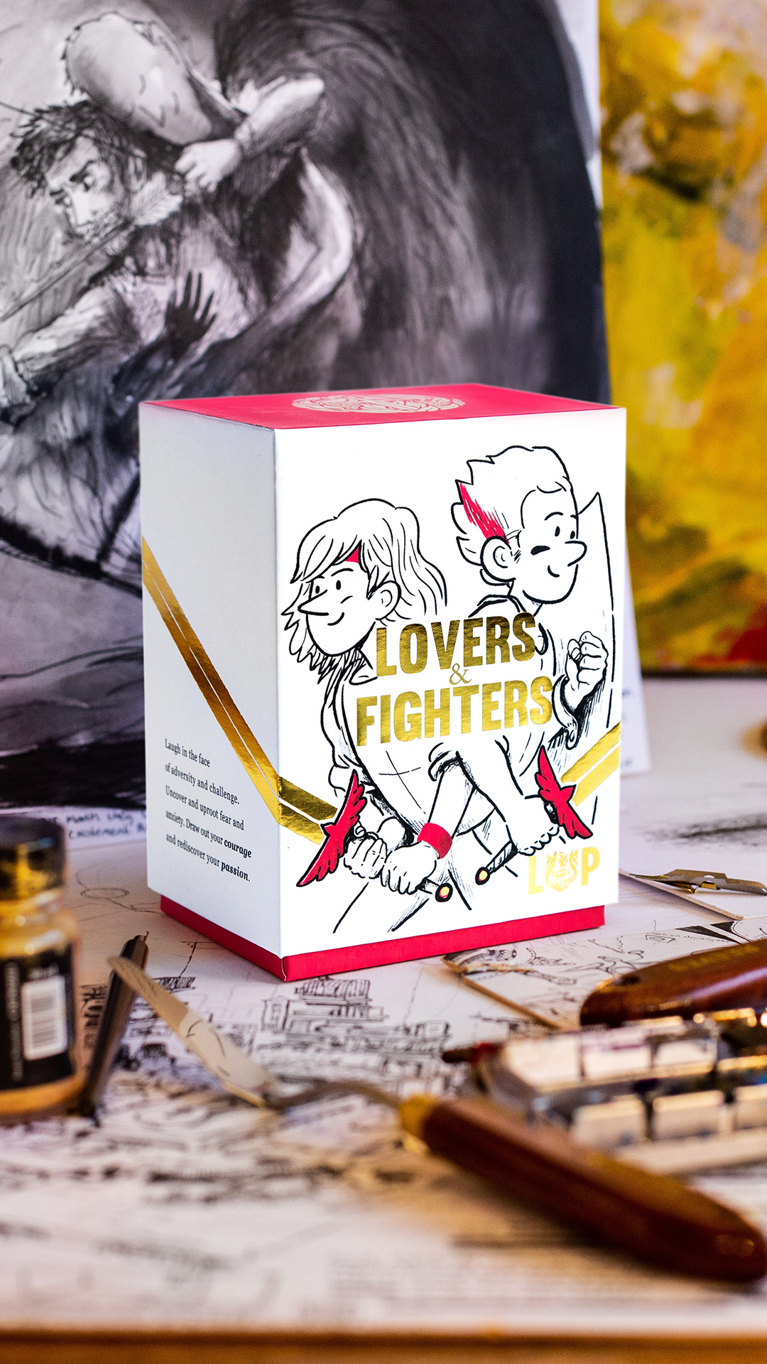 Living & Powerful 'Lovers & Fighters' boxed card set, gold foiled, on a desk with hand drawn artwork behind it